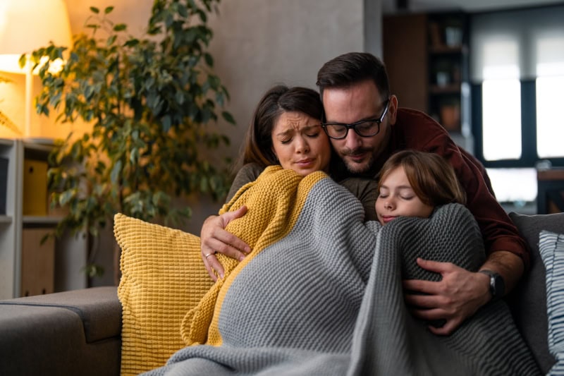 Why Isn’t My Furnace Heating My Entire Home? Photo of a family of three, freezing in their home huddled under a blanket to keep warm.